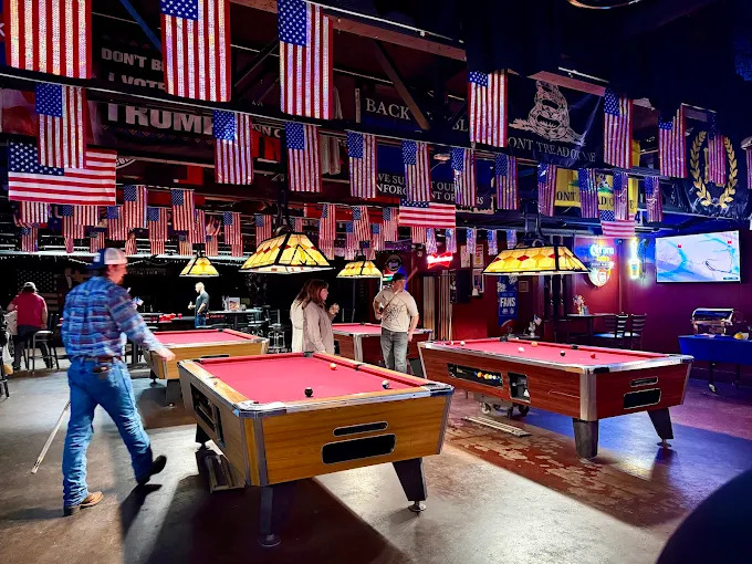Slingshots pool hall interior — multiple pool tables under warm pendant lights, flags and neon signs on the walls
